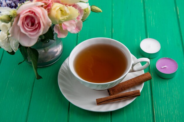 Vista lateral de la taza de té con canela en platillo y velas con flores sobre fondo verde