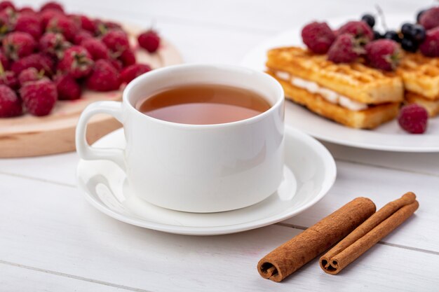 Vista lateral de la taza de té con cerezas blancas canela y waffles dulces con frambuesas sobre una superficie blanca