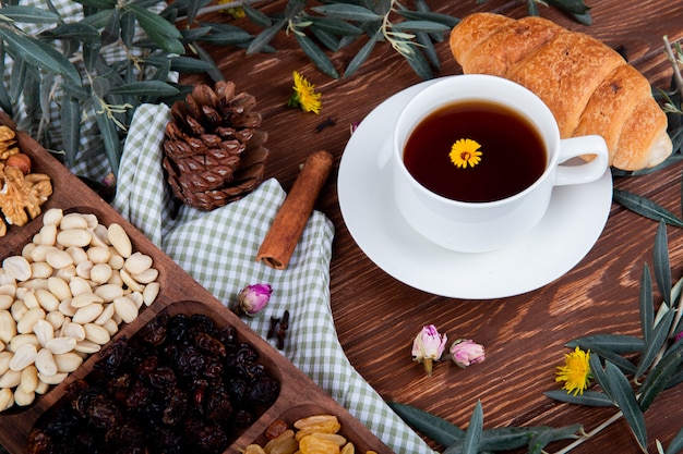 Vista lateral de una taza de té con croissant, nueces mixtas con frutos secos y dientes de león dispersos en madera