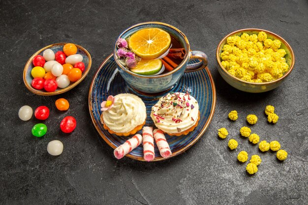 Vista lateral de una taza de té, cuencos de dulces coloridos junto al plato de un apetitoso cupcake con crema y una taza de té en la mesa