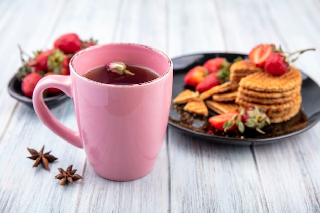 Vista lateral de la taza de té con flores y galletas de gofres con fresas en placas sobre superficie de madera