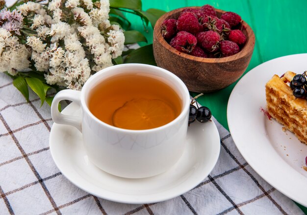 Vista lateral de una taza de té con frambuesas y flores sobre una toalla a cuadros