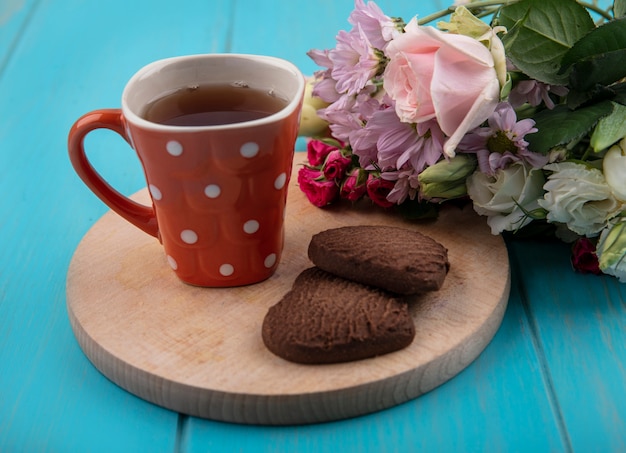 Vista lateral de la taza de té y galletas en forma de corazón en la tabla de cortar con flores sobre fondo azul.