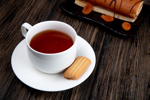 Vista lateral de una taza de té con galletas y pastel de rollo en una bandeja sobre superficie de madera rústica