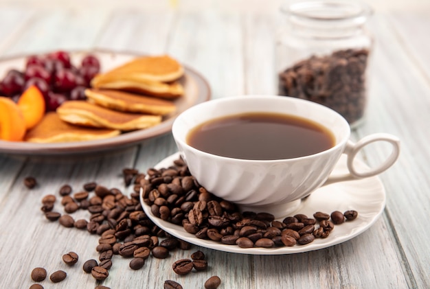 Vista lateral de la taza de té y granos de café en el platillo con plato de panqueques y cerezas y rodajas de albaricoque con jarra de granos de café sobre fondo de madera