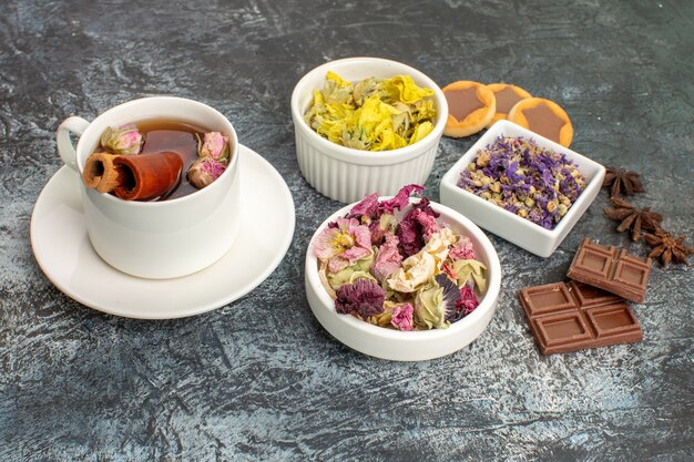 Vista lateral de una taza de té de hierbas con cuencos de flores secas y dulces sobre suelo gris