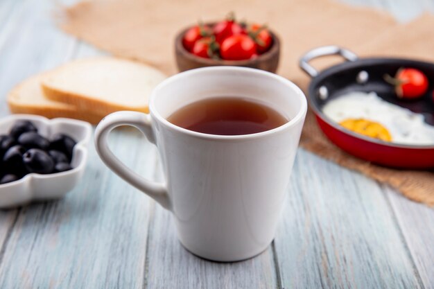 Vista lateral de la taza de té con huevo frito de tomate sobre cilicio y rebanadas de pan de aceitunas negras sobre superficie de madera