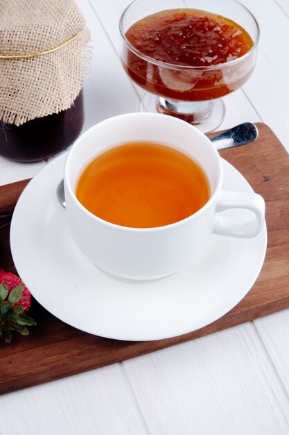 Vista lateral de una taza de té con mermelada de fresa en un florero de vidrio sobre tabla de madera en blanco