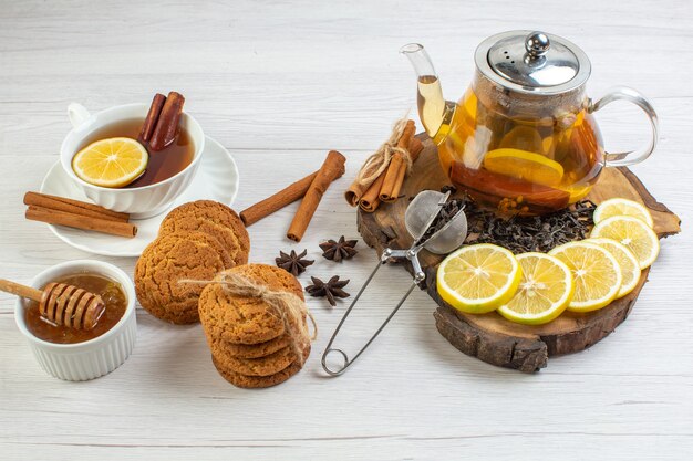 Vista lateral de una taza de té negro galletas miel y té de hierbas en una olla de vidrio y limones de canela limón picados en una bandeja de madera sobre fondo blanco.