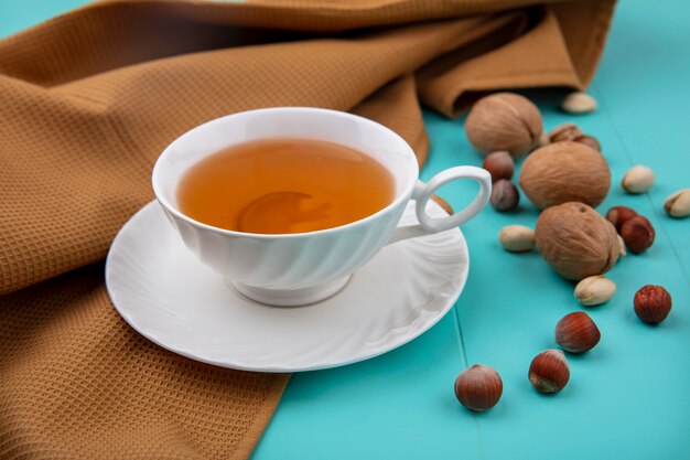 Vista lateral de la taza de té con nueces avellanas con pistachos con una toalla marrón sobre una superficie turquesa