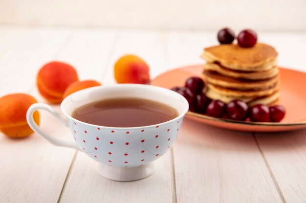 Vista lateral de la taza de té con plato de panqueques y cerezas y albaricoques sobre fondo de madera