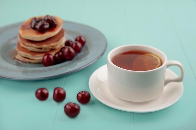 Vista lateral de la taza de té en plato y panqueques con cerezas en placa y sobre fondo azul.