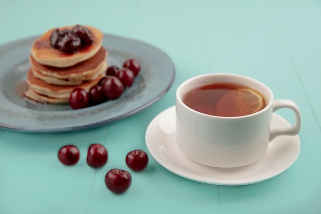Vista lateral de la taza de té en plato y panqueques con cerezas en placa y sobre fondo azul.