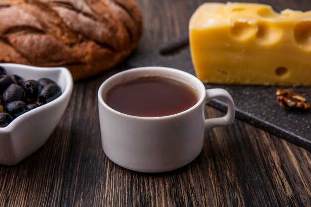 Vista lateral de una taza de té con queso maasdam en un soporte con aceitunas y pan negro sobre la mesa