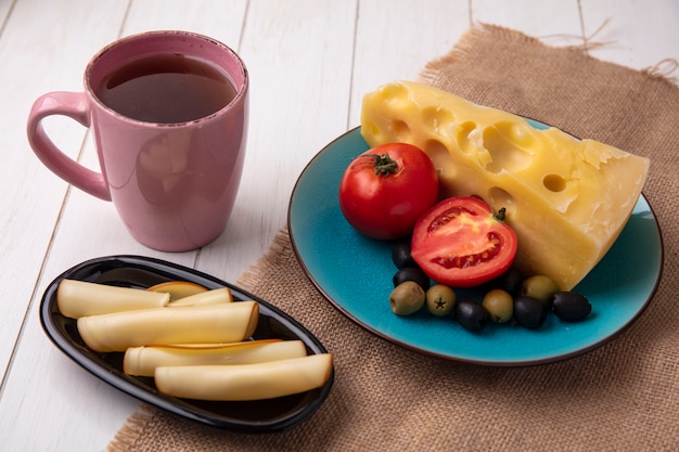 Vista lateral de una taza de té con quesos, tomate, aceitunas en un plato y ahumado sobre un fondo blanco.