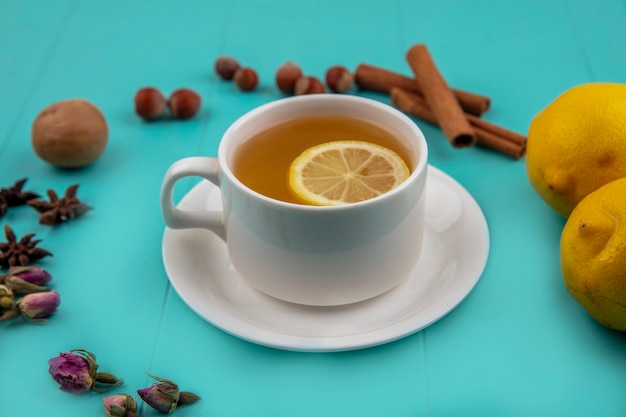 Vista lateral de la taza de té con una rodaja de limón y canela con nueces nueces limones y flores sobre fondo azul.