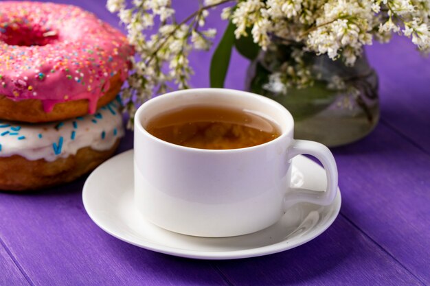 Vista lateral de la taza de té con rosquillas y flores sobre una superficie de color púrpura brillante