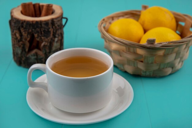 Vista lateral de la taza de té con un tazón de canela y una canasta de limones sobre fondo azul.