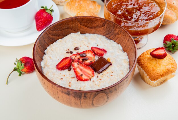 Vista lateral del tazón de avena con queso cottage chocolate y fresas con mermelada de magdalenas rollos de té en mesa blanca