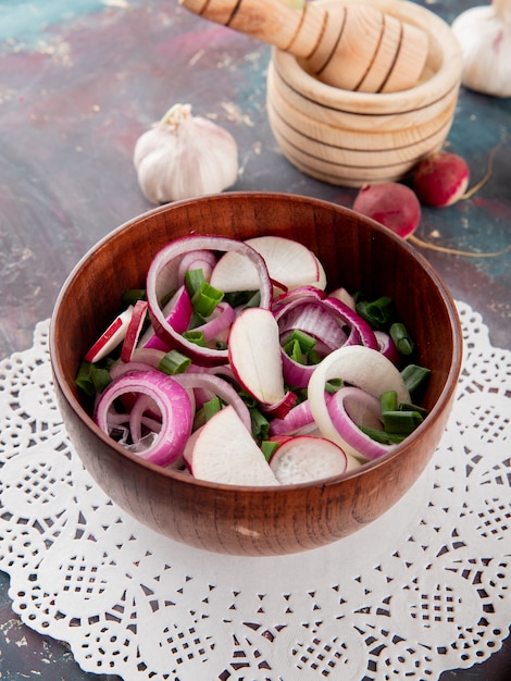 Vista lateral del tazón de ensalada de verduras con cebolla rábano y cebolleta en papel tapete con ajo sobre fondo marrón