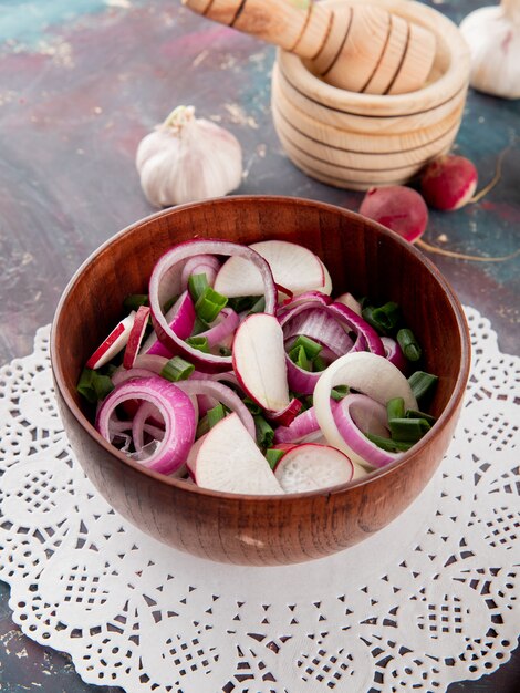 Vista lateral del tazón de ensalada de verduras con cebolla rábano y cebolleta en papel tapete con ajo sobre fondo marrón