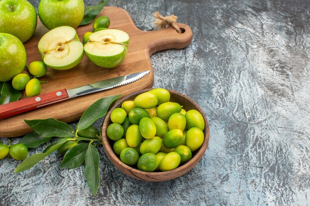 Vista lateral del tazón de frutas de cítricos, manzanas verdes y un cuchillo en la tabla de cortar
