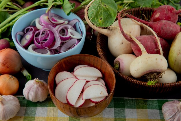 Vista lateral de tazones y canasta de verduras como cebolla de rábano y ajo en la superficie de la tela