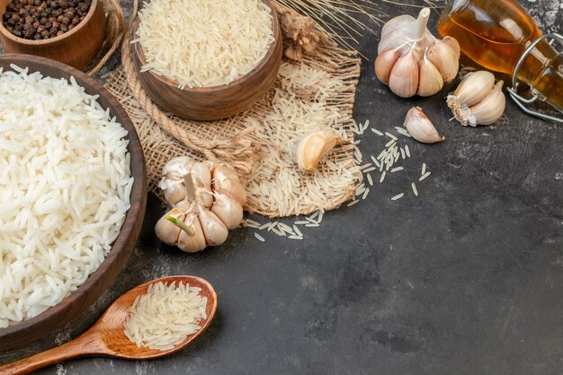 Vista lateral de tazones largos perfectos de arroz marrón sobre una toalla de color desnudo, una botella de aceite caída, ajos, pimientos sobre una mesa oscura