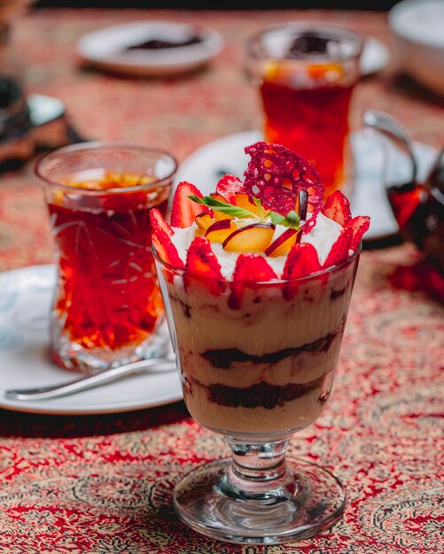 Vista lateral tiramisú de postre en un vaso con rodajas de fresas y manzana y un vaso de té