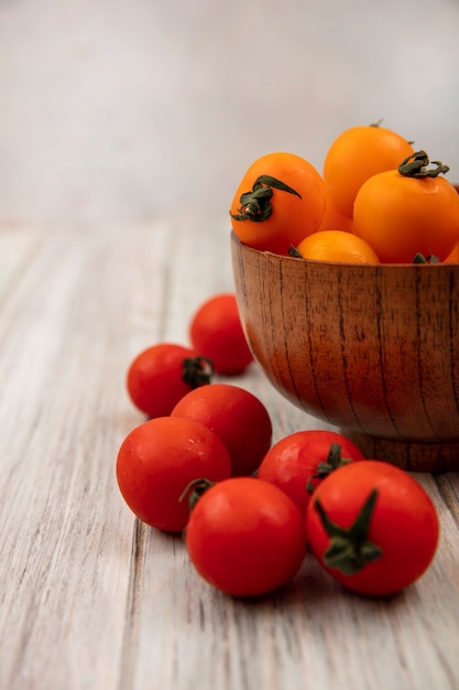 Vista lateral de tomates naranjas en un cuenco de madera con tomates rojos aislado sobre una superficie de madera gris