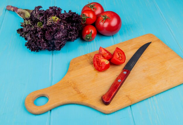 Vista lateral de tomates en rodajas con cuchillo en la tabla de cortar y albahaca con tomates enteros sobre superficie azul