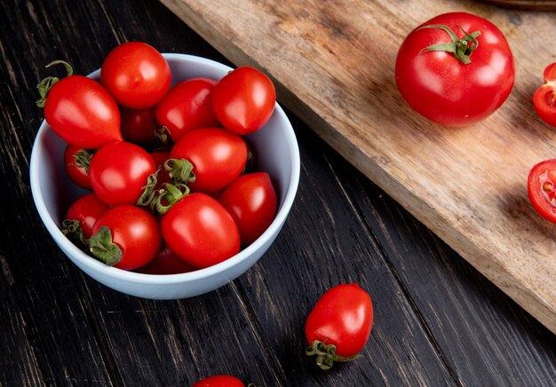 Vista lateral de tomates en un tazón y otros en tabla de cortar en mesa de madera