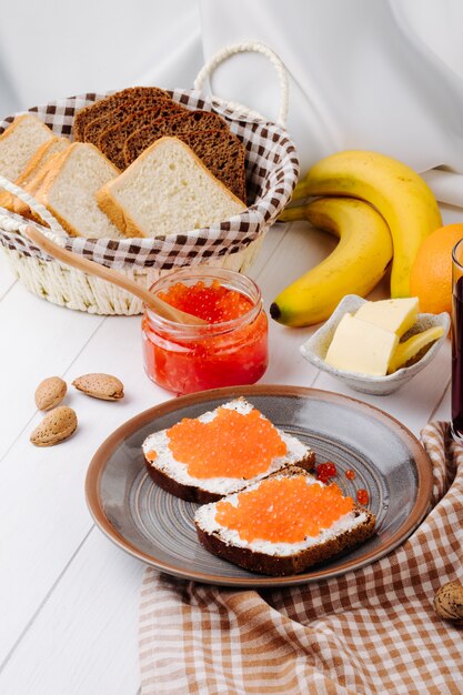 Vista lateral tostadas de caviar rojo pan de centeno con queso cottage caviar rojo mantequilla pan blanco naranja plátano y almendras sobre la mesa