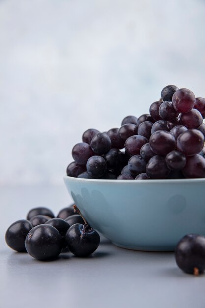 Vista lateral de uvas negras frescas en un recipiente azul sobre un fondo gris