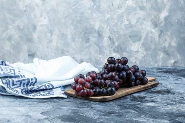 Vista lateral de uvas negras con tabla de cortar sobre yeso sucio y fondo de toalla de cocina. horizontal