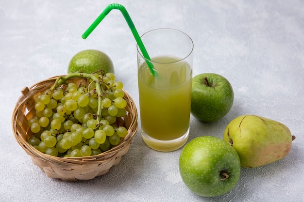 Vista lateral de uvas verdes en una canasta con pera manzanas verdes y jugo de manzana con una pajita verde en un vaso sobre un fondo blanco.