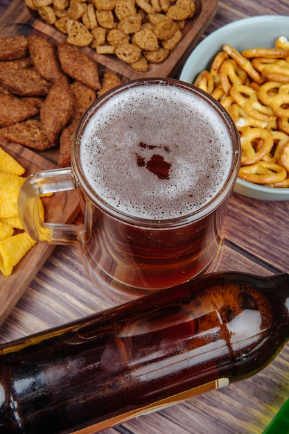 Vista lateral de variados aperitivos de cerveza, galletas saladas, papas fritas y mini pretzels con una jarra de cerveza en rústico