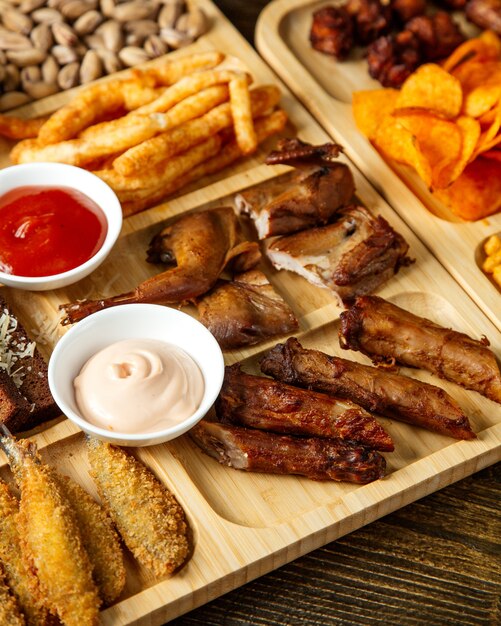 Vista lateral de una variedad de bocadillos de cerveza como guails a la parrilla, papas fritas, pistachos y papas fritas con salsas sobre una tabla de madera