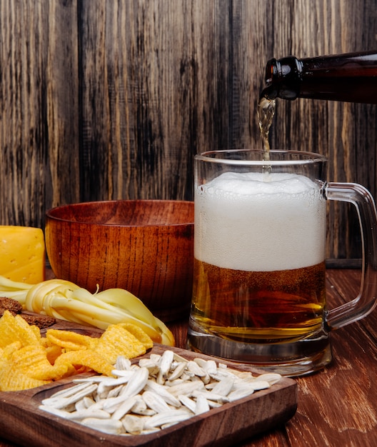 Vista lateral de varios bocadillos de cerveza salada en una bandeja de madera y verter cerveza en una taza sobre madera rústica