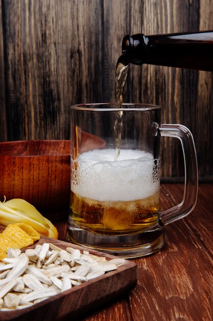 Vista lateral de varios bocadillos de cerveza salada en una bandeja de madera y verter cerveza en una taza sobre madera rústica