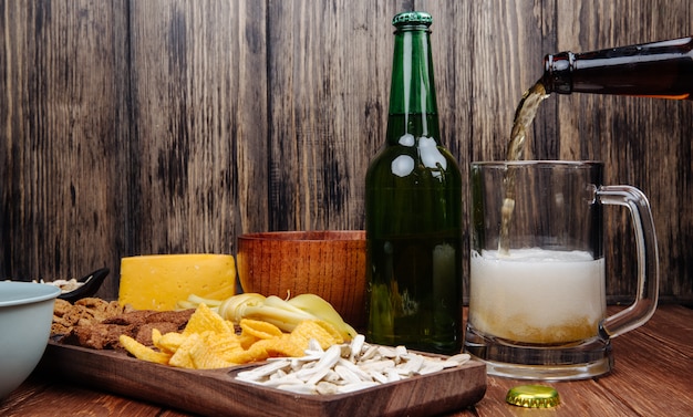 Vista lateral de varios bocadillos de cerveza salada en una bandeja de madera y verter cerveza en una taza sobre madera rústica