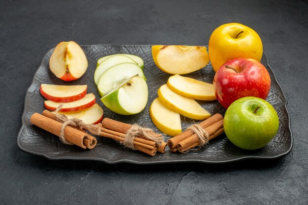 Vista lateral de varios tipos de manzanas frescas en rodajas y enteras en una bandeja negra y limones canela en el lado derecho sobre un fondo oscuro