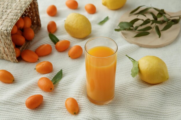 Vista lateral del vaso de jugo de naranja con hojas de limones y kumquats derramándose de la canasta sobre fondo de tela blanca