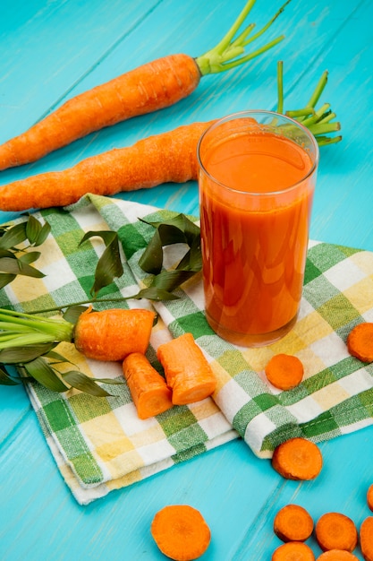 Foto gratuita vista lateral del vaso de jugo de zanahoria con zanahorias cortadas y en rodajas sobre tela decorada con hojas en la mesa azul