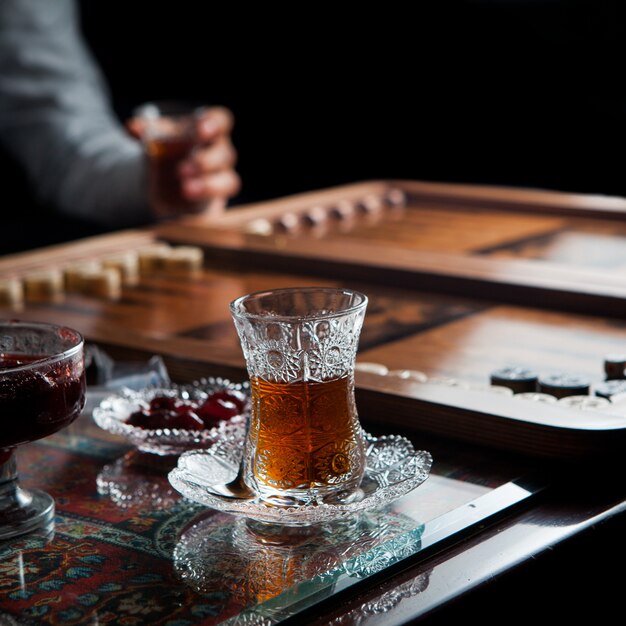 Vista lateral vaso de té con backgammon y mano humana y mermelada en zócalo en la mesa de la alfombra