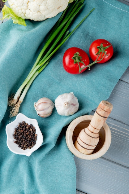 Vista lateral de verduras como cebolleta tomate ajo con pimienta negra sobre tela y trituradora de ajo sobre fondo de madera