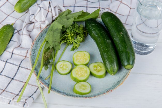 Vista lateral de verduras como pepino entero y en rodajas de espinacas y cilantro con pepinos sobre tela y agua de desintoxicación en mesa de madera