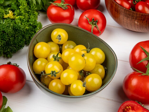 Vista lateral de verduras como tomate cilantro con tazón de tomates amarillos en mesa de madera