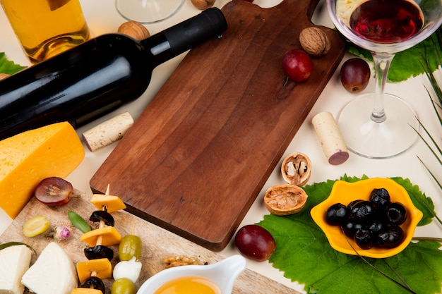 Vista lateral de vino tinto con diferentes tipos de queso mantequilla de oliva en tabla de cortar y corchos de nuez vino blanco sobre fondo blanco.