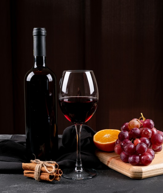 Vista lateral vino tinto con uva, naranja en tabla de cortar de madera en vertical oscuro